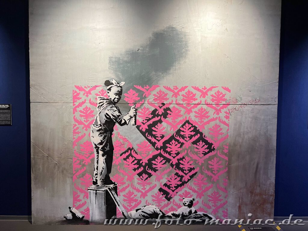 Banksy im Kunstkraftwerk - Mädchen auf einer Kiste übersprüht schwarzes Hakenkreuz mit pinkfarbenen Mustern